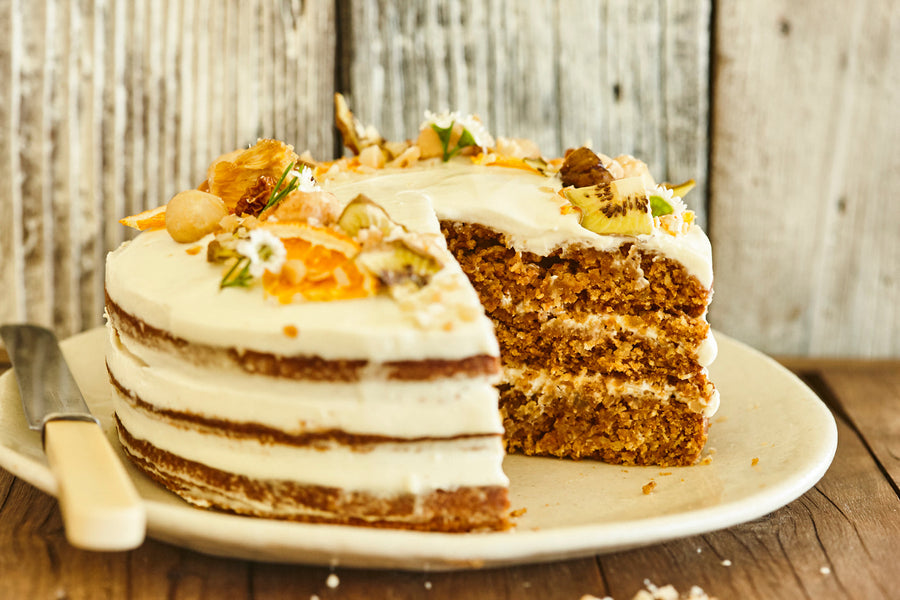 A slice removed from a frosted macadamia carrot and ginger spice cake topped with nuts and fruit.