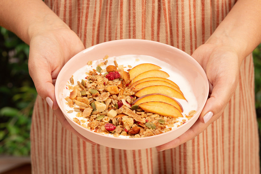 A person holding a pink bowl of granola with yogurt, peach slices, and nuts.