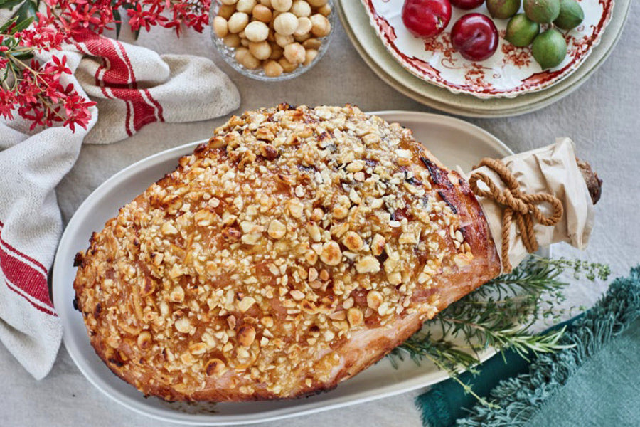 Roasted glazed ham coated in crushed macadamia nuts on a white platter with rosemary garnish