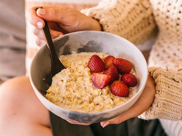 Woman in sweater holds a spoon and a bowl of Brookfarm porridge with sliced strawberries
